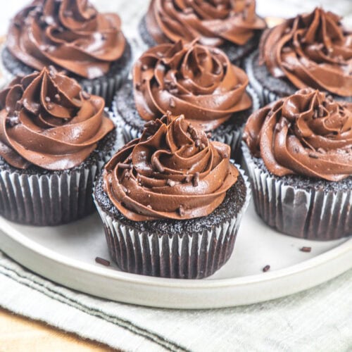 7 eggless chocolate cupcakes with chocolate buttercream frosting on a plate.