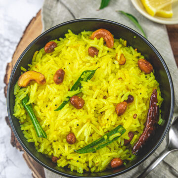 Lemon rice topped with peanuts, cashews, curry leaves with a spoon and lemon slices on the side