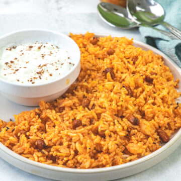 A plate with vagharelo bhaat and a bowl of yogurt with pickle bottles in the back.