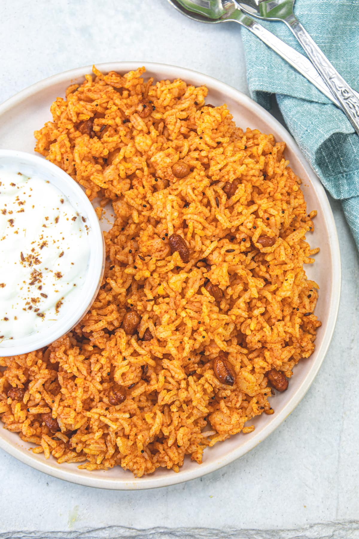 Vagharelo bhaat in a plate with a bowl of yogurt and 2 spoons on the side with napkin.