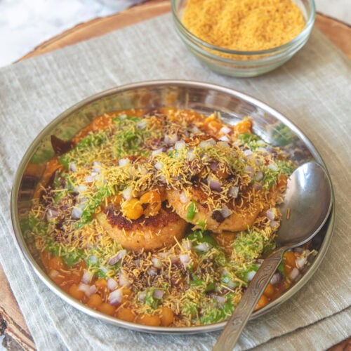 A plate of ragda patties chaat with a spoon, a bowl of sev, onion and chutney in the back.