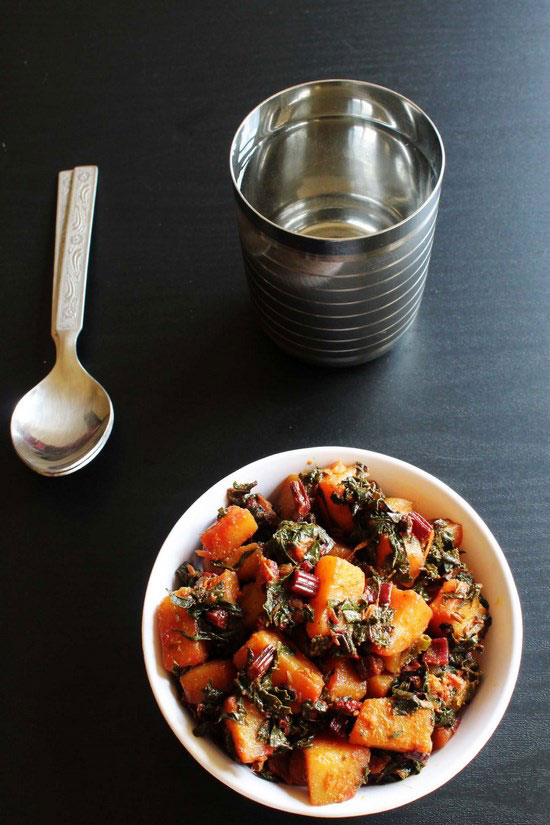 Aloo beet greens sabzi in a bowl with spoons and a glass of water in the back.