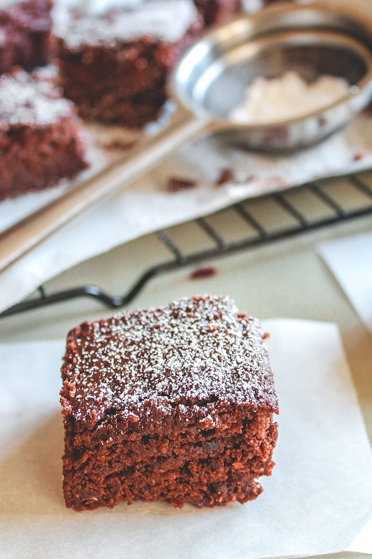 A piece of brownie without eggs in a parchment paper with more pieces in the back.