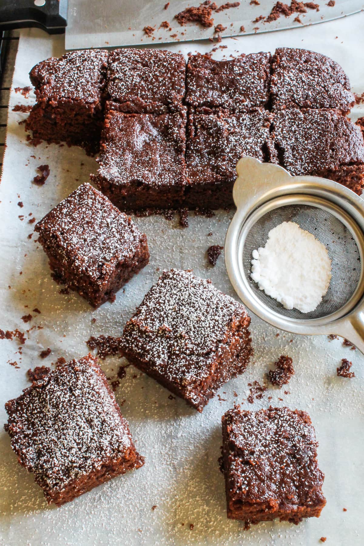 Eggless brownies pieces on a cooling rack with parchment paper and a strainer with powdered sugar.