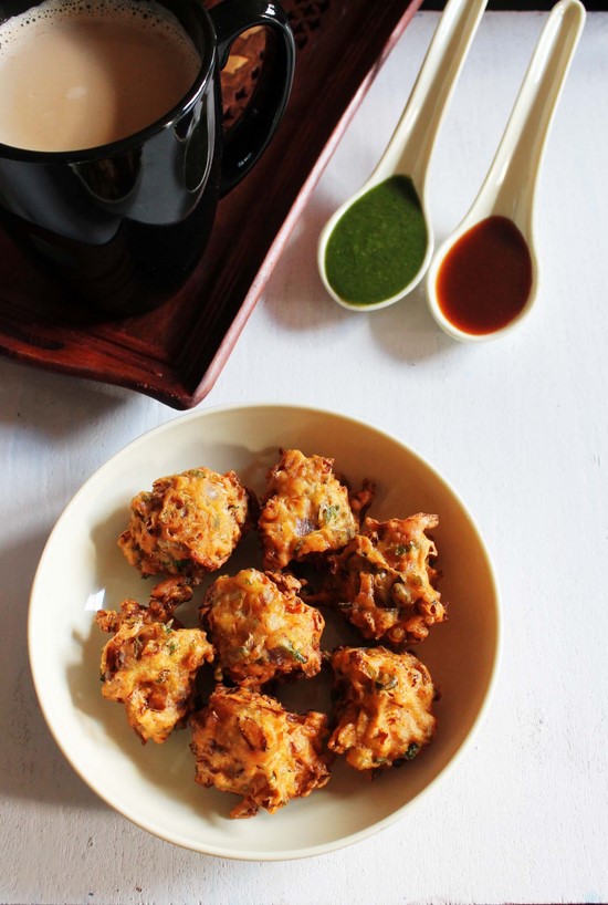 Cabbage pakoda in a plate with 2 chutneys and a cup of tea in the back.