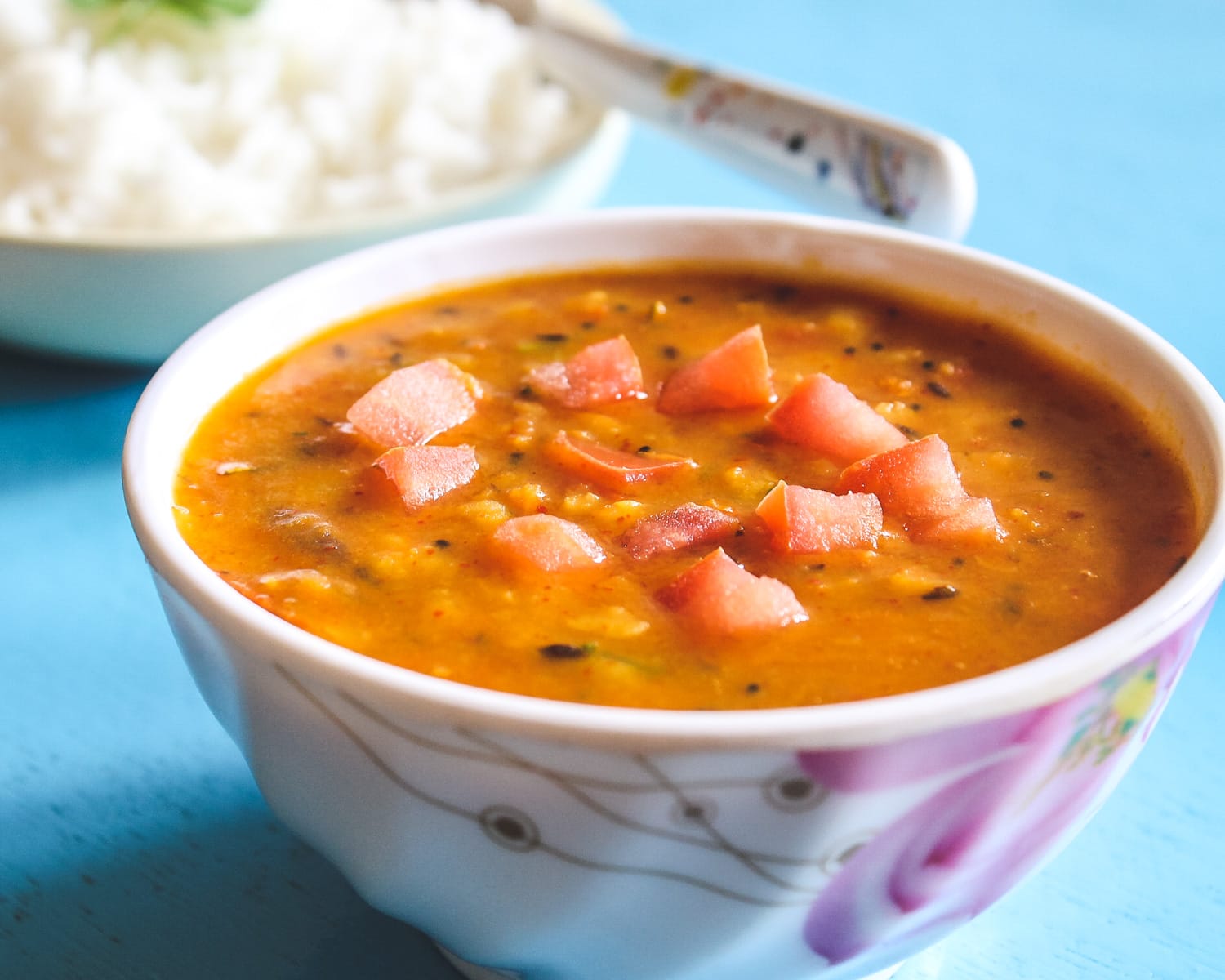 A bowl of tomato dal garnished with tomatoes and rice in the back.