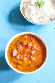 A bowl of tomato dal with a plate of rice in the back.