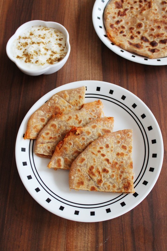 Mooli paratha triangles in a plate with spiced yogurt bowl and more paratha in the back.