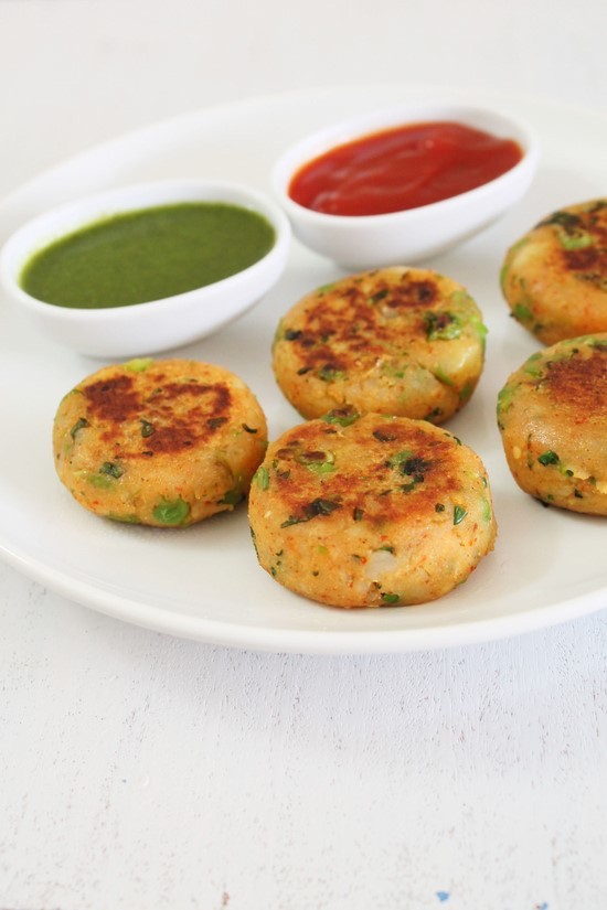 5 Aloo tikki in a white plate with small bowls of ketchup and green chutney.