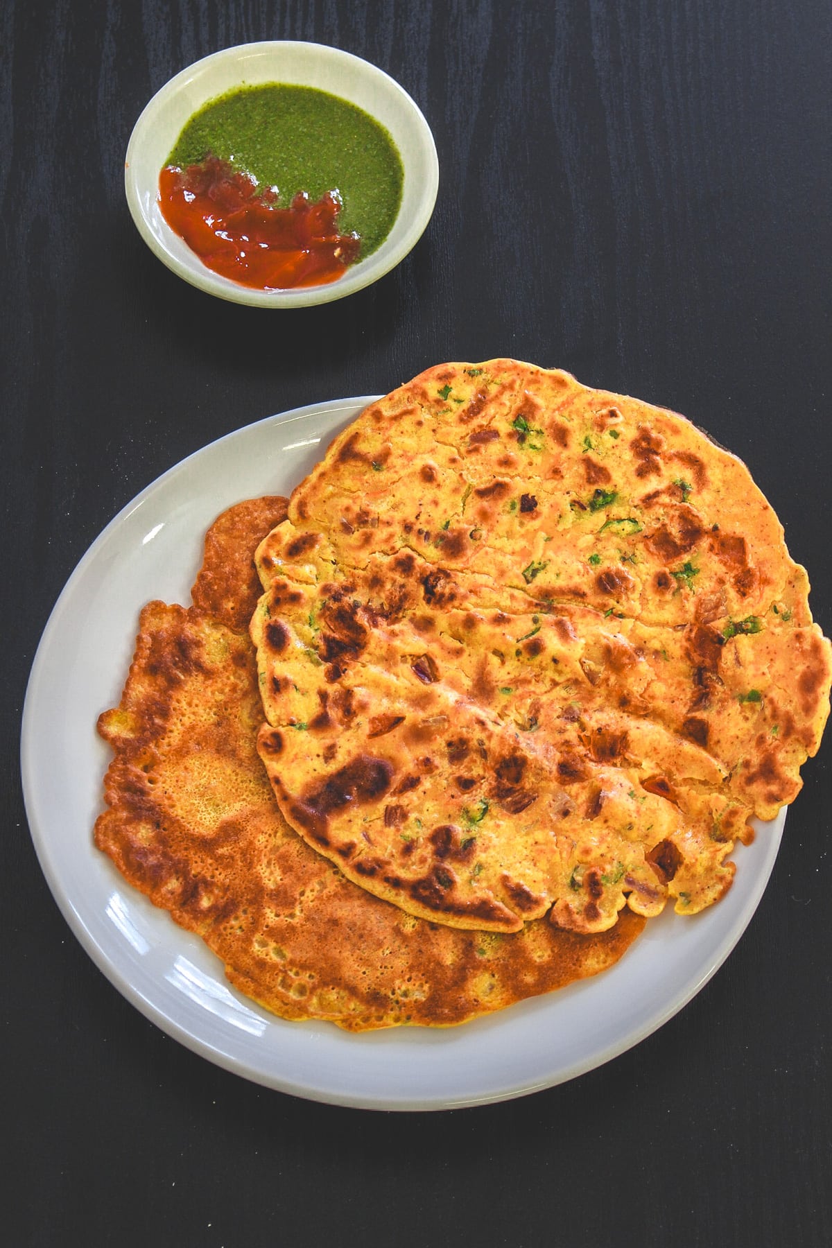 2 besan chilla on the plate with green chutney and ketchup on the side.