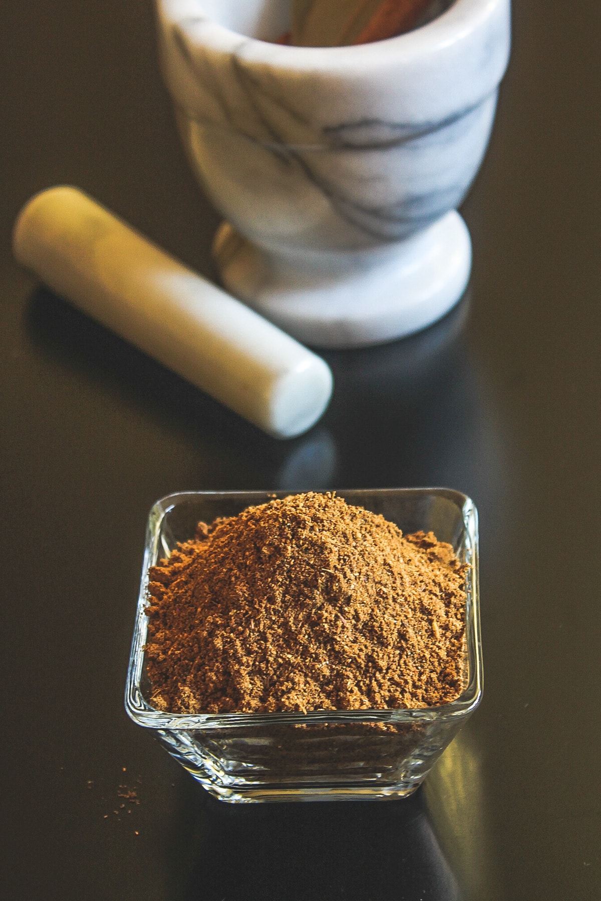 Homemade garam masala in a square bowl with mortar pestle in back.