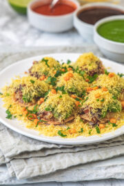 A plate of sev puri with 3 bowls of chutney in the back.