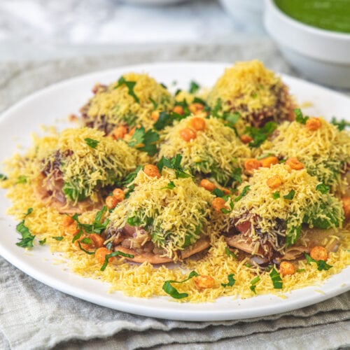 A plate of sev puri with 3 bowls of chutney in the back.