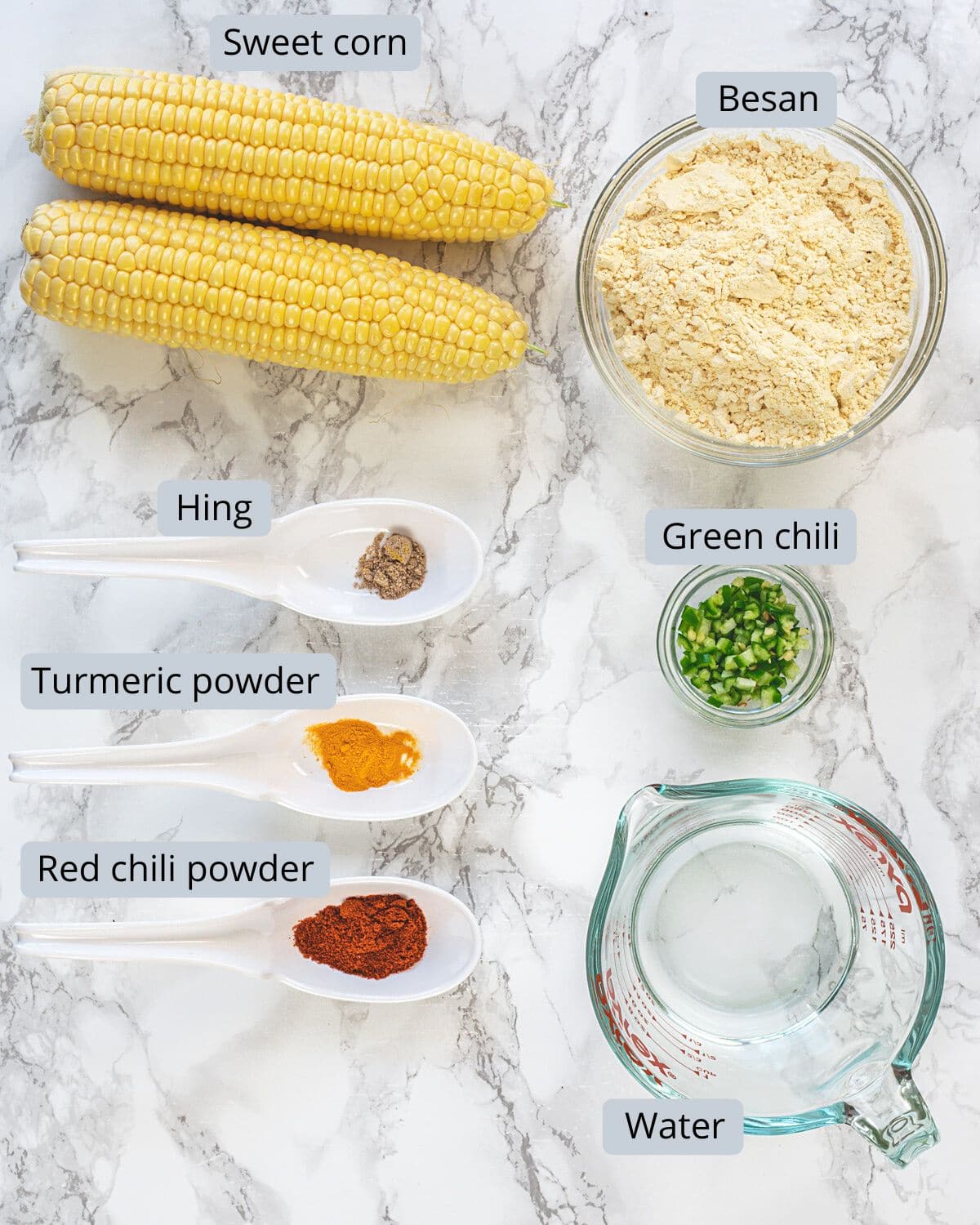 Corn pakoda ingredients in bowls and spoons on marble surface with labels.