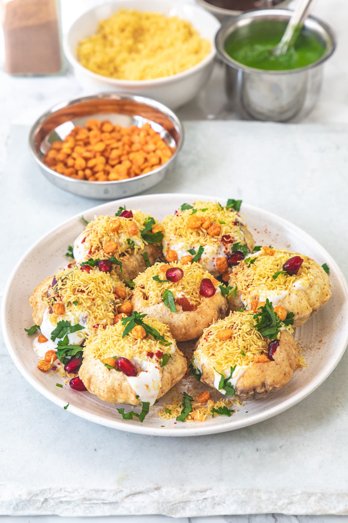 Dahi puri plate on a marble board with bowls of chana dal, green chutney and sev in the back.