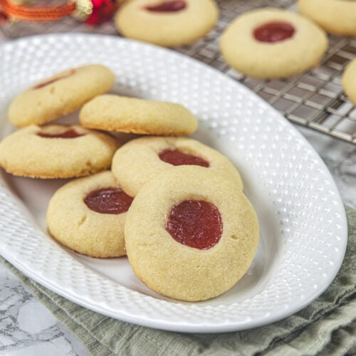 Shortbread thumbprint cookies in a plate and a few more in the back on a cooling rack.