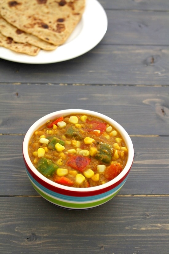 Corn capsicum masala served in a bowl with a plate of paratha in the back.