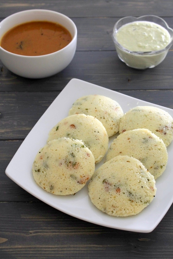 6 rava idli in a square plate with a bowl of sambar and a small bowl of chutney in the back.