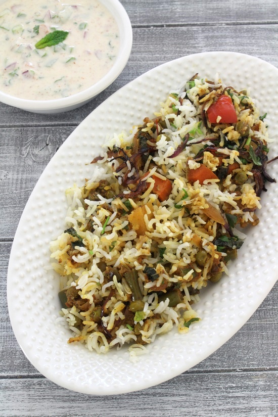 Veg biryani in a white oval plate with vegetable raita in the back.