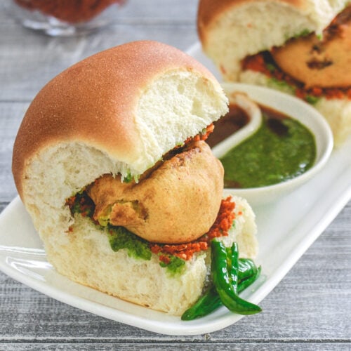 2 vada pav in a plate with fried chili, green chutney and tamarind chutney with garlic chutney in the back.