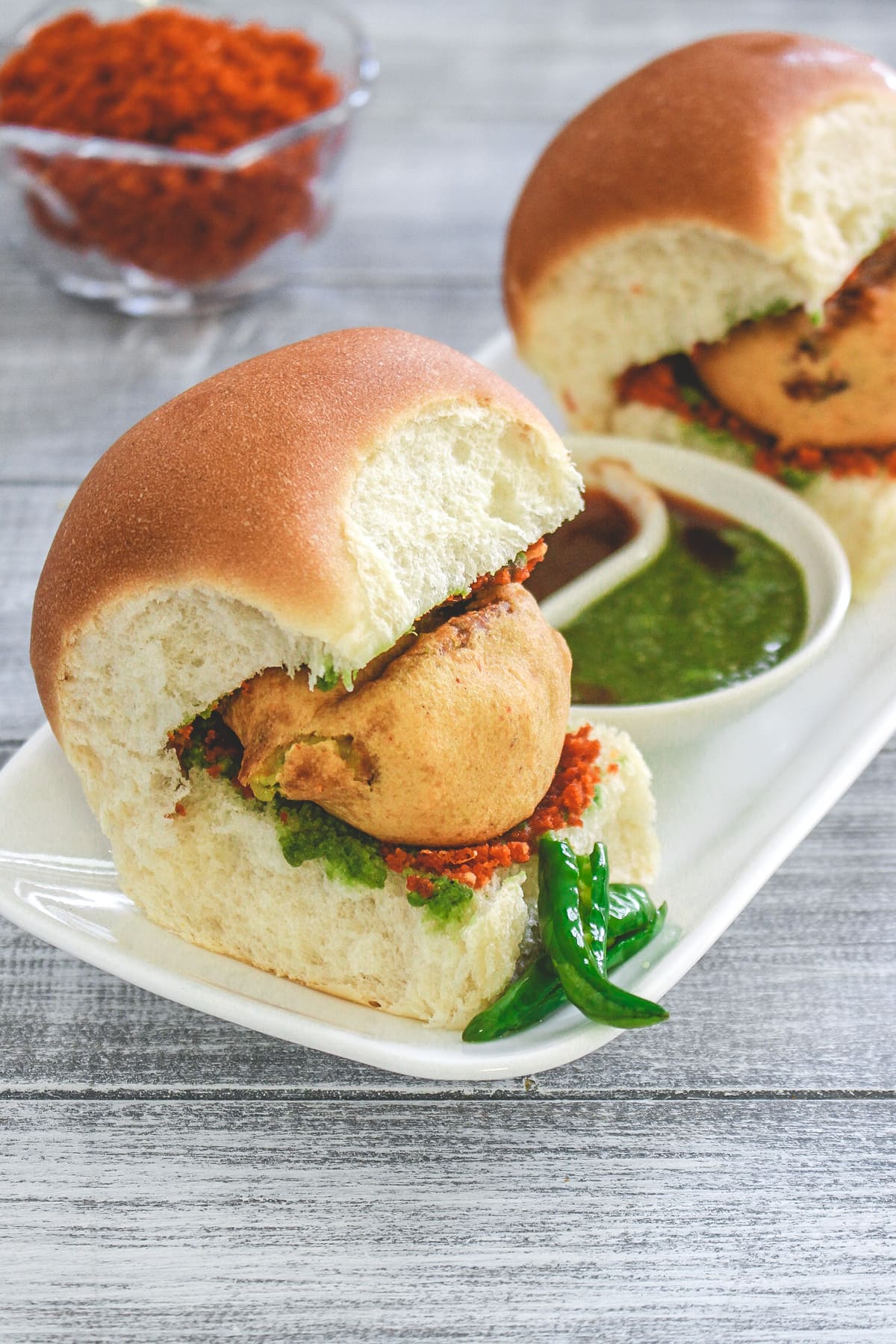 2 vada pav in a plate with fried chili, green chutney and tamarind chutney with garlic chutney in the back.