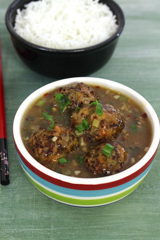 Veg manchurian served in a bowl and garnished with spring onion greens and a bowl of rice in the back.