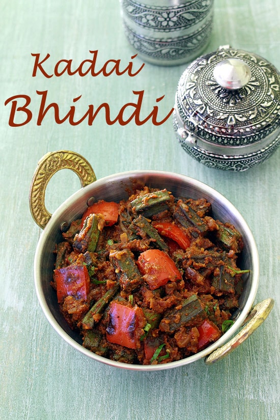 Kadai Bhindi served in a serving kadai with steel bowl and glass in the back.