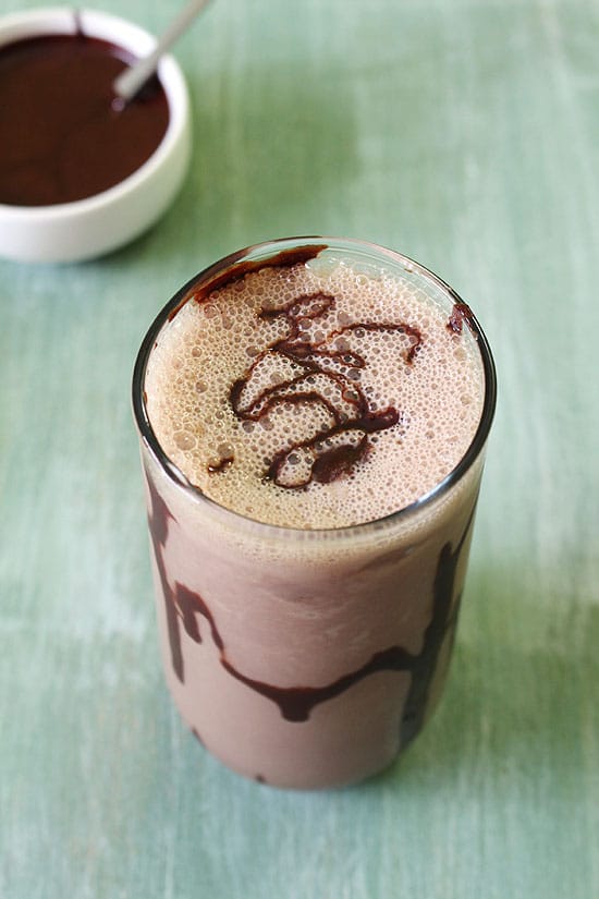 Chocolate milkshake with a drizzle of chocolate syrup on top and a bowl of chocolate sauce in the back.