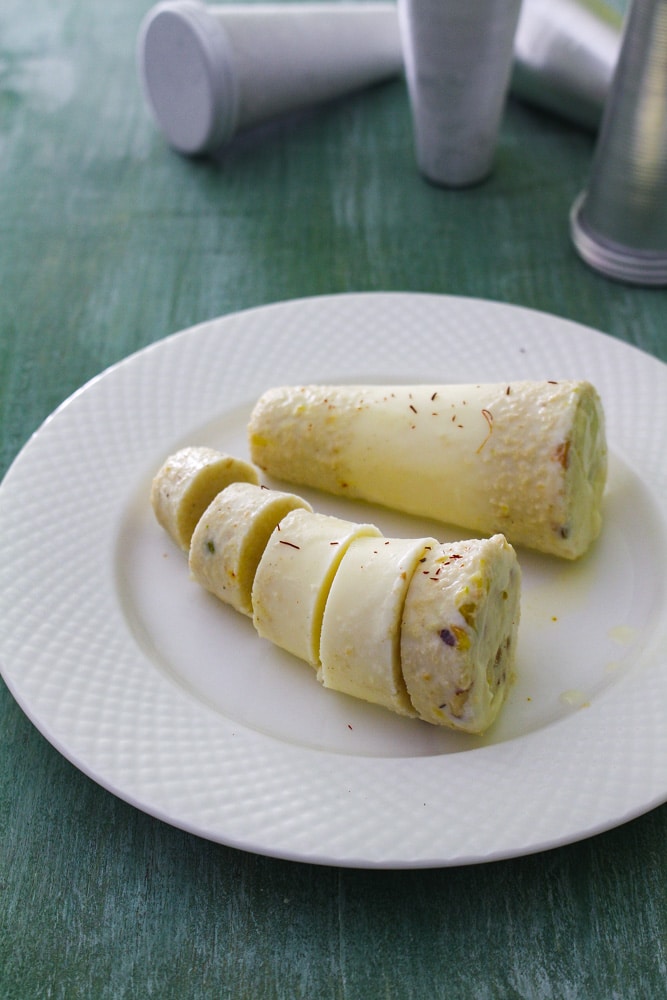 1 whole kulfi and 1 slices of kulfi in a plate with kulfi moulds in the back.