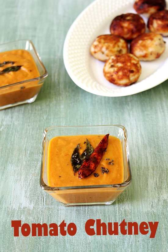 Tomato chutney garnished with tadka and another bowl of chutney and a plate of paniyaram in the back.