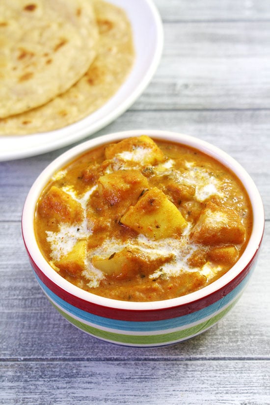 Aloo paneer garnished with a drizzle of cream and a plate of paratha in the back.