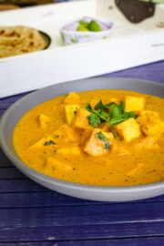 Paneer butter masala in a bowl with garnish of cilantro, paratha and onion in the back in a tray