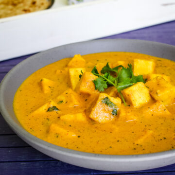 Paneer butter masala in a bowl with garnish of cilantro, paratha and onion in the back in a tray