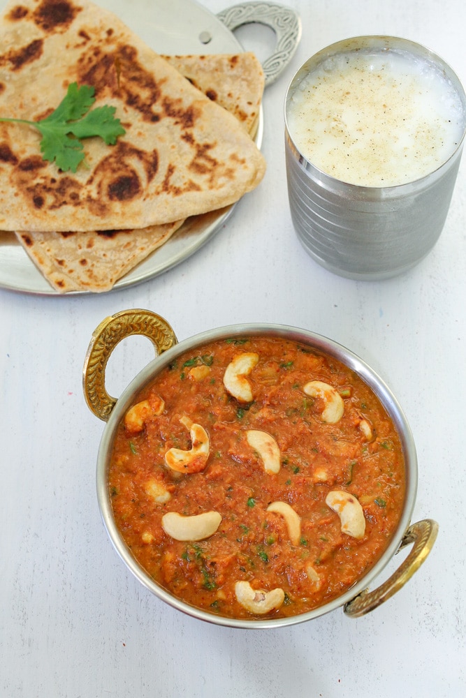 Kaju curry in a serving kadai with paratha and chaas in the back.