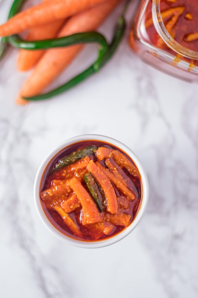 Carrot pickle in a bowl with carrots and green chilies with a jar of pickle in the back.