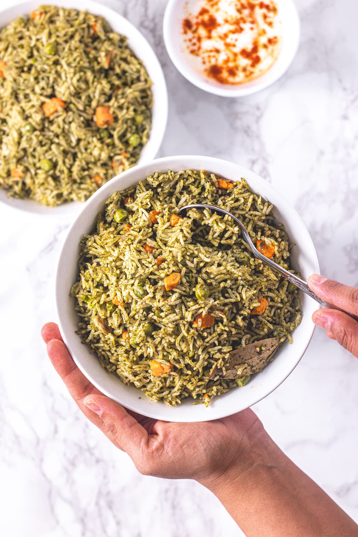 Taking a spoonful of spinach pulao from the bowl, another pulao bowl and yogurt bowl in the back.