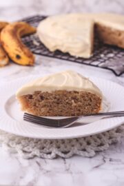 A slice of eggless banana cake in a plate with fork, few bananas and remaining cake in the back.