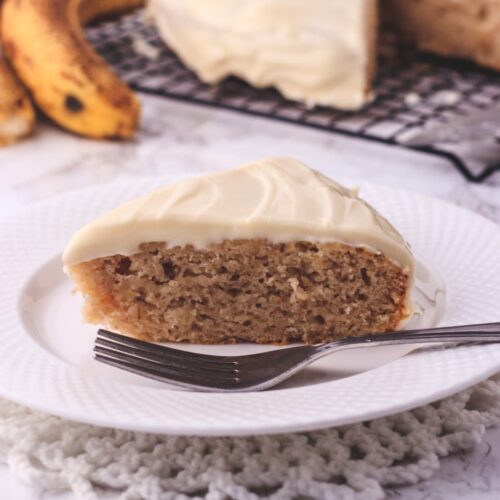 A slice of eggless banana cake in a plate with fork, few bananas and remaining cake in the back.