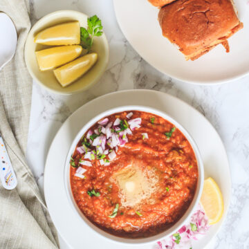 top view of pav bhaji in a bowl, buns, onion and lemon wedges on side