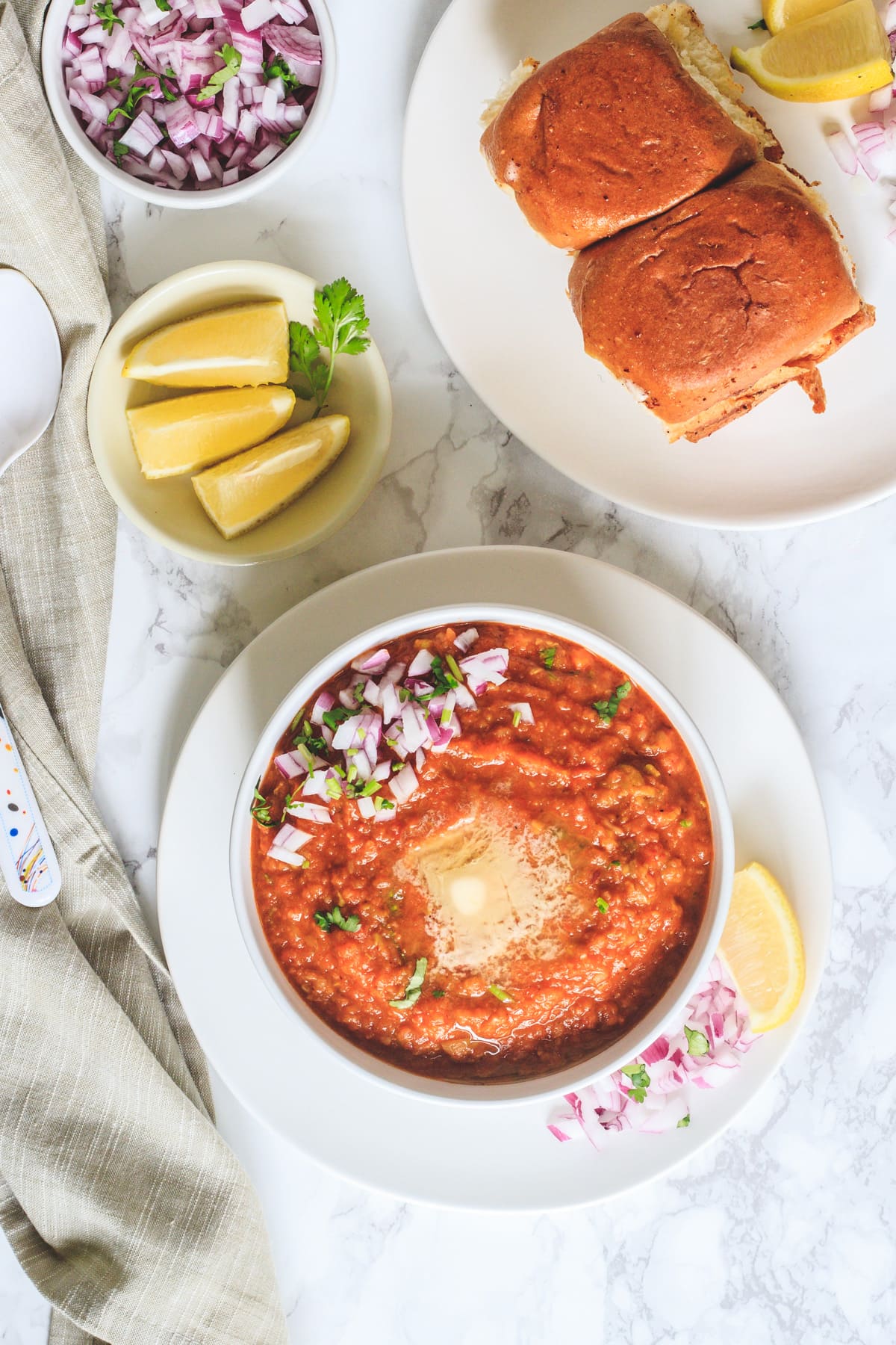 top view of pav bhaji in a bowl, buns, onion and lemon wedges on side