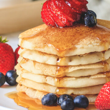 close up of stacked pancakes in a plate with garnish of berries and pouring maple syrup