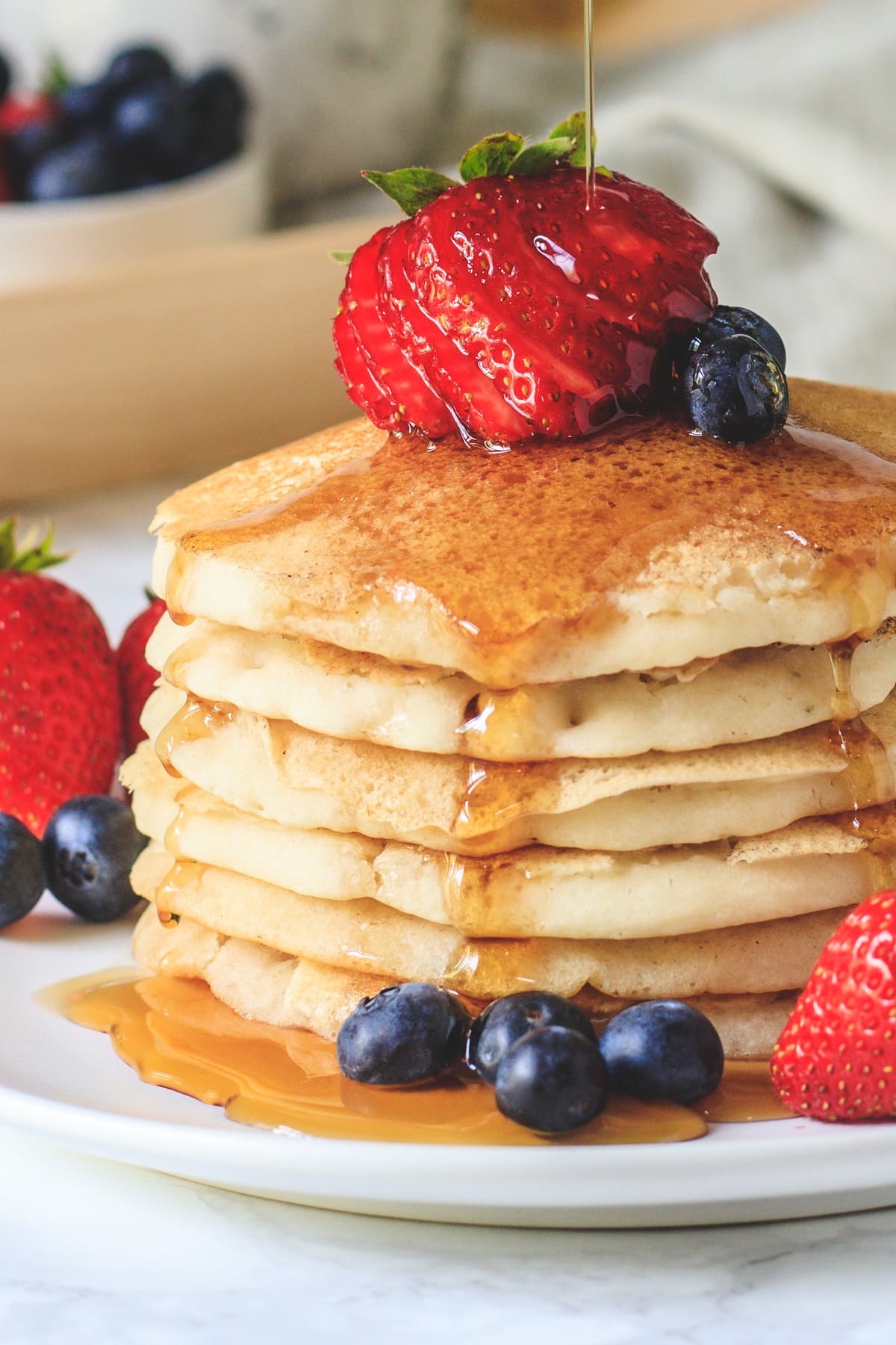 close up of stacked pancakes in a plate with garnish of berries and pouring maple syrup