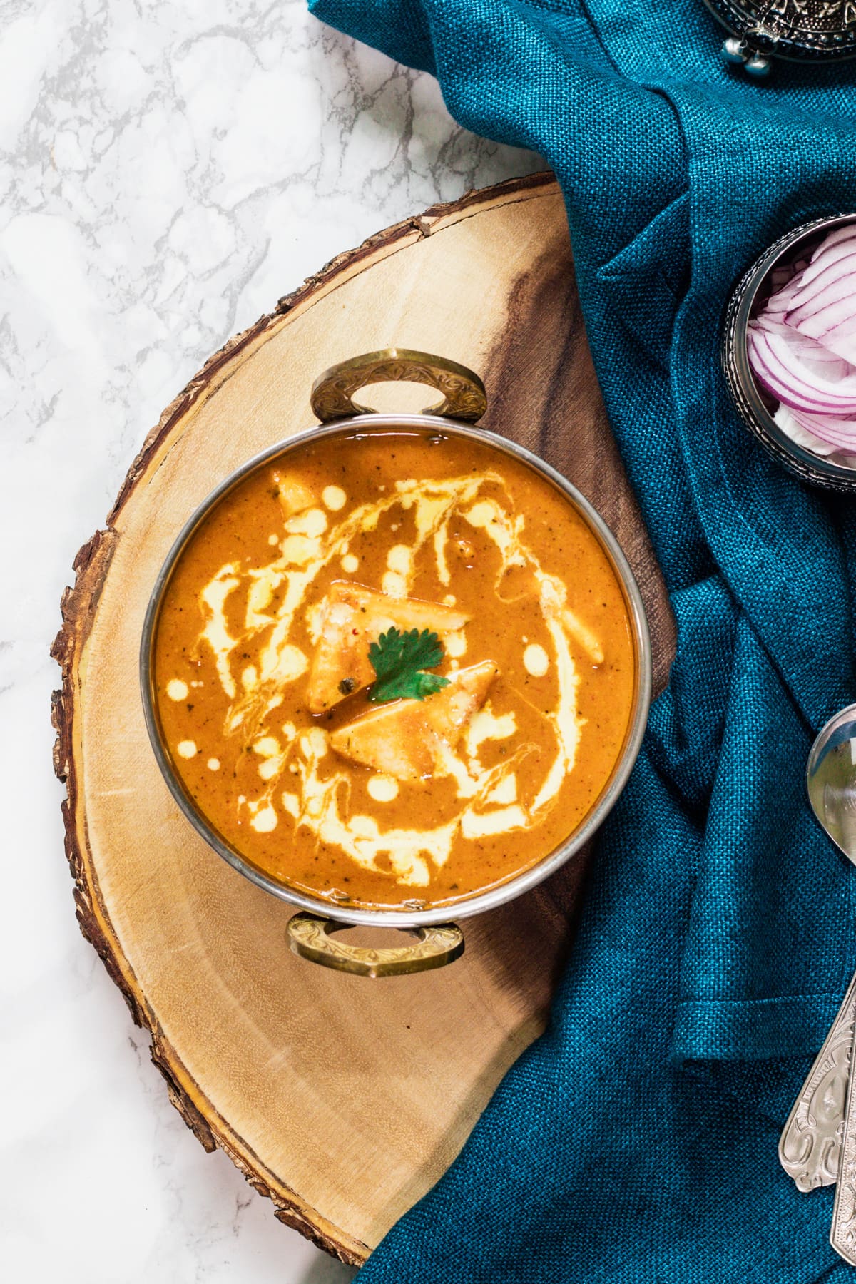 Shahi paneer with a garnish of cream and cilantro in a kadai on a wooden board with napkin, onion, spoons