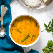 A bowl of gujarati dal with side of papad & rice with napkin and spoons on the side with few cilantro & curry leaves