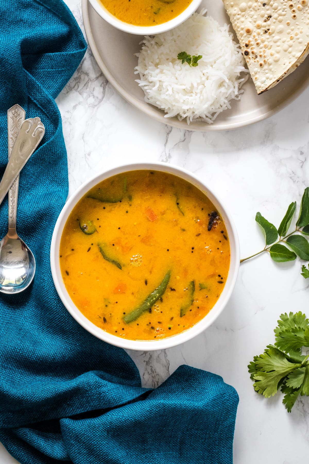 A bowl of gujarati dal with side of papad & rice with napkin and spoons on the side with few cilantro & curry leaves