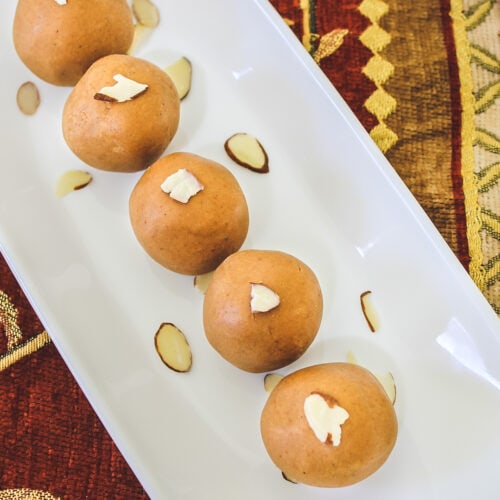 5 besan ladoo garnished with almonds arranged on rectangle plate with decorative napkin underneath