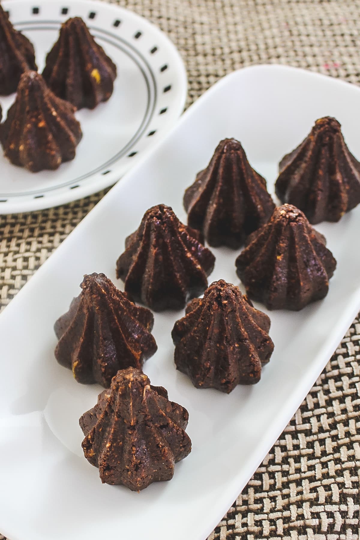 Top view of 6 chocolate modak in white rectangle plate with 3 of them on round plate in back