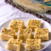 7 pieces of coconut burfi in a plate with few pieces in a pan in the back