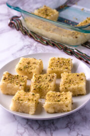7 pieces of coconut burfi in a plate with few pieces in a pan in the back