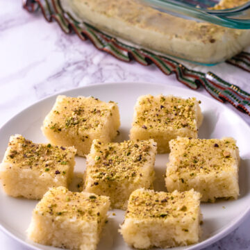 7 pieces of coconut burfi in a plate with few pieces in a pan in the back